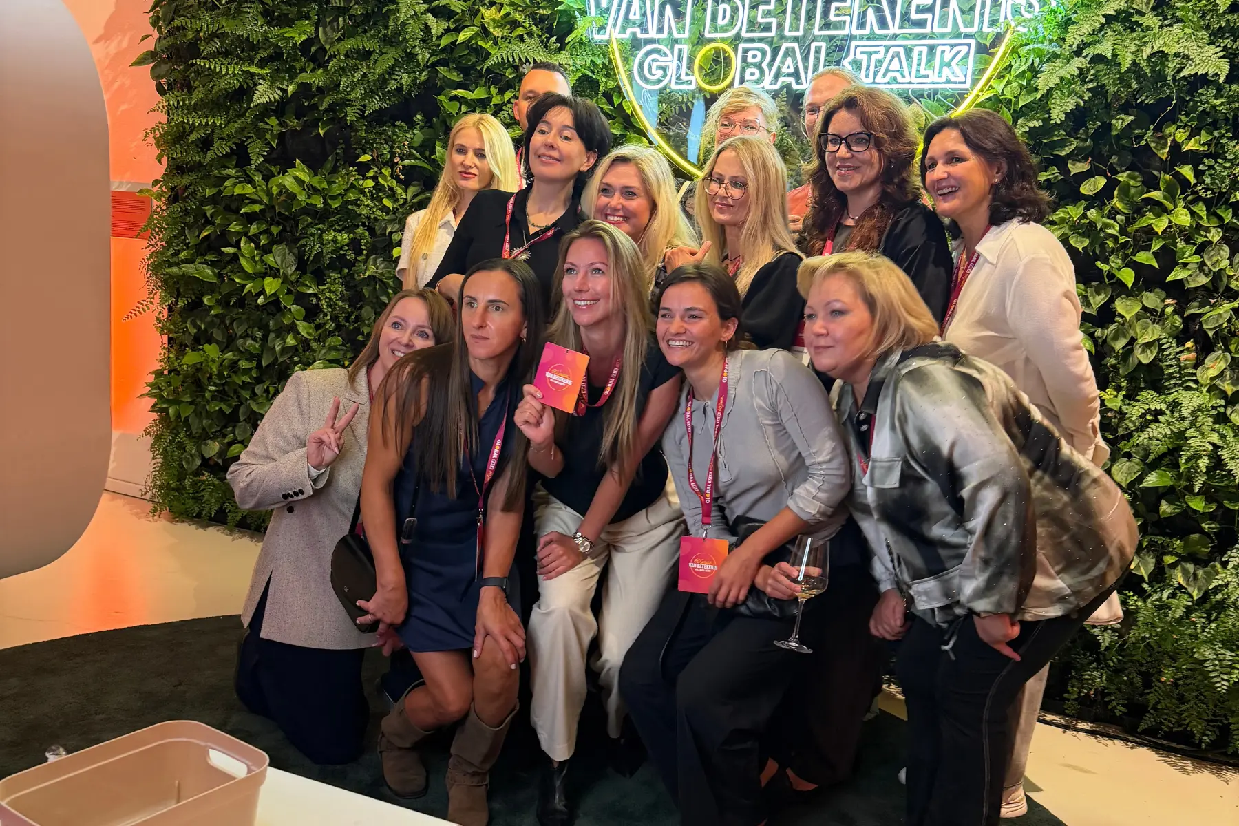 Groep collega's met badges poseert samen voor een groene wand met neonlogo