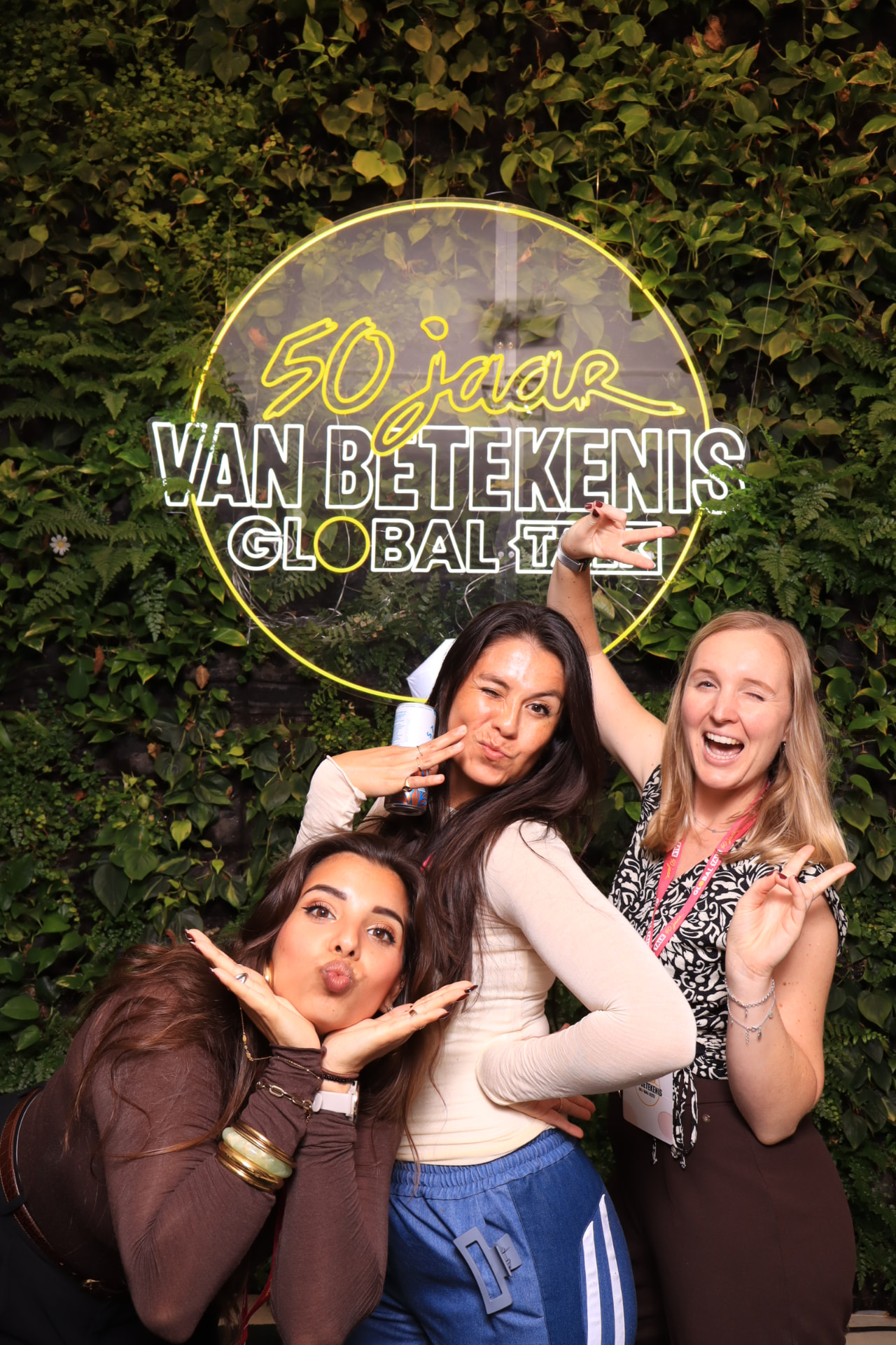 Drie vrouwen poseren speels onder een neonbord voor een groene plantenwand