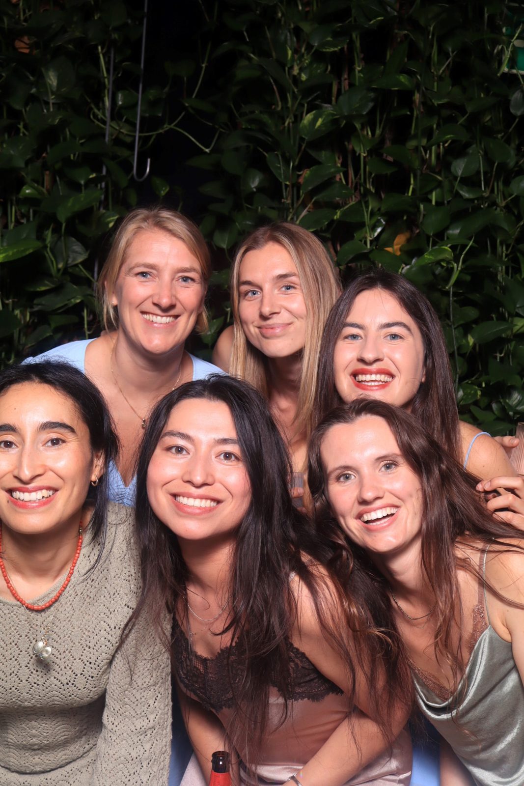 Zes lachende vrouwen staan dicht bij elkaar voor een groene plantenwand