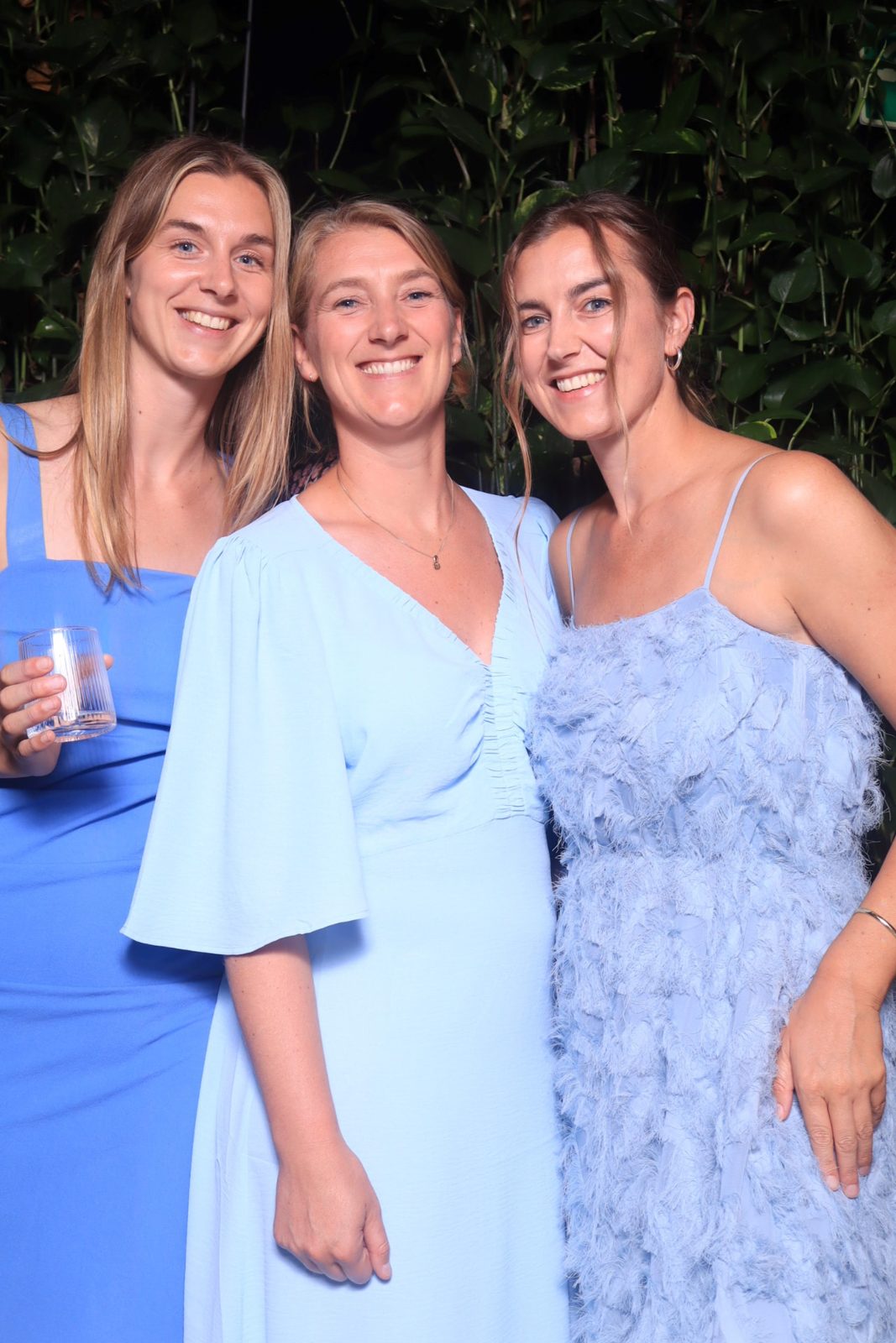 Drie vrouwen in blauwe jurken lachen samen voor een groene plantenwand